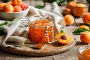 Selbstgemachte Aprikosenmarmelade in einem Einmachglas, umgeben von frischen und überreifen Aprikosen auf einem Holzbrett
