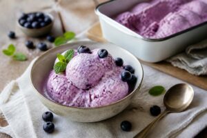 Cremiges Heidelbeer-Joghurt-Eis ohne Eismaschine in einer weißen Schale, mit frischen Blaubeeren garniert, auf einem Holztisch