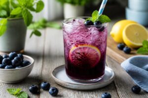 Zuckerfreie Heidelbeer-Limonade im Glas mit Eiswürfeln, frischen Blaubeeren und Minzblättern auf hellem Hintergrund