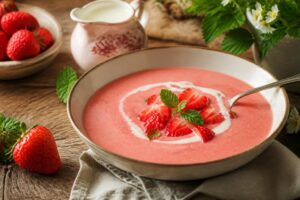 Kalte Erdbeersuppe nach Omas Art in einer weißen Suppenschüssel, garniert mit Sahnehaube und frischen Minzblättern, umgeben von reifen Erdbeeren auf einem Holztisch