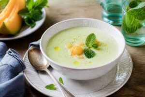 Kalte Melonensuppe mit Minze in einem klaren Glasschälchen, garniert mit frischen Minzblättern und Melonenwürfeln, von oben fotografiert auf hellem Holzuntergrund