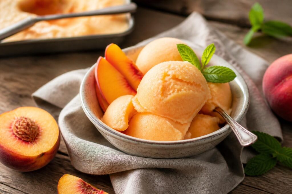Cremiges Pfirsich-Sorbet ohne Eismaschine in einer Glasschale, garniert mit frischer Minze und Pfirsichscheiben auf einem Holztisch