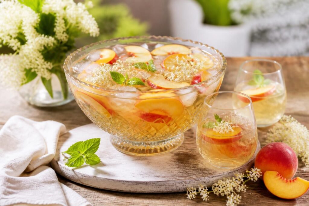 Alkoholfreie Sommerbowle mit frischen Pfirsichscheiben, Holunderblütensirup und Eiswürfeln in einem Glaskaraffe, dekoriert mit Minzzweigen auf einem Holztisch im Freien